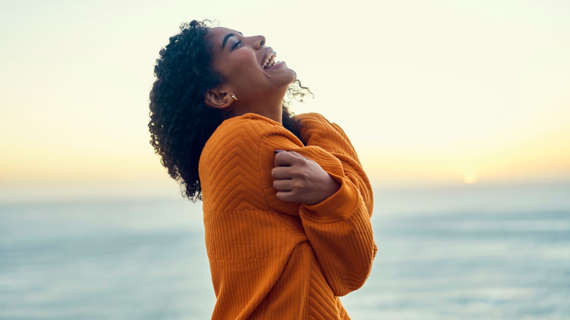 Mujer con un suéter naranja sonriendo junto al océano al atardecer.